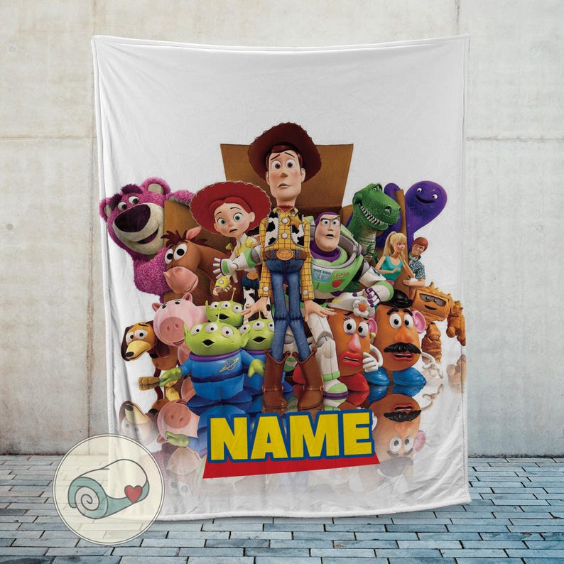 May include: White blanket featuring a colourful group of Toy Story characters, including Woody, Buzz Lightyear, and Mr. Potato Head. The word "NAME" is printed in yellow with a red border at the bottom. The blanket is displayed against a light grey wall.