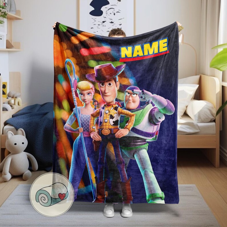 Puede incluir: Una manta de felpa con los personajes Woody, Buzz Lightyear y Bo Peep de Toy Story. La manta tiene un fondo azul oscuro con la palabra "NAME" en amarillo y rojo. La manta est&aacute; siendo sostenida por una persona.