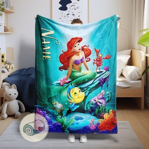 Puede incluir: Una manta turquesa con una colorida ilustraci&oacute;n de Ariel de La Sirenita. Ariel, con cabello rojo y un top de concha p&uacute;rpura, est&aacute; sentada en una roca con Flounder, Sebasti&aacute;n y otros personajes. La palabra "NAME" est&aacute; impresa verticalmente.