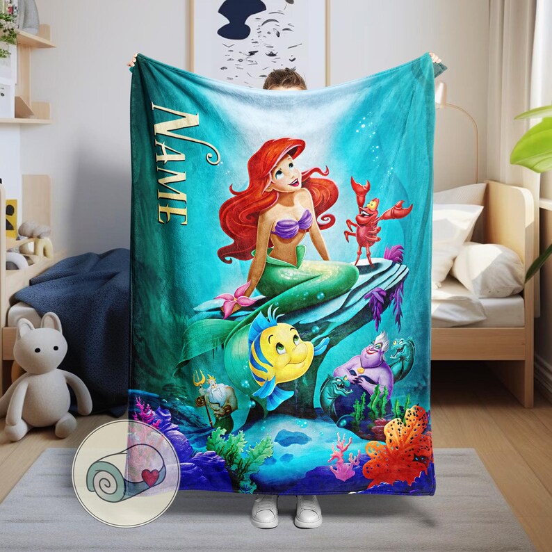 Puede incluir: Una manta turquesa con una colorida ilustraci&oacute;n de Ariel de La Sirenita. Ariel, con cabello rojo y un top de concha p&uacute;rpura, est&aacute; sentada en una roca con Flounder, Sebasti&aacute;n y otros personajes. La palabra "NAME" est&aacute; impresa verticalmente.