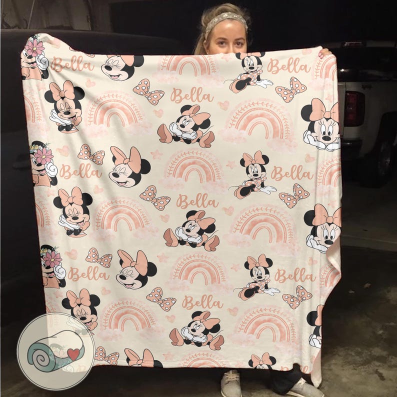 May include: A cream-colored blanket featuring a repeating pattern of Minnie Mouse, bows, rainbows, and the name "Bella" in a soft pink hue. The blanket is held up by a person.