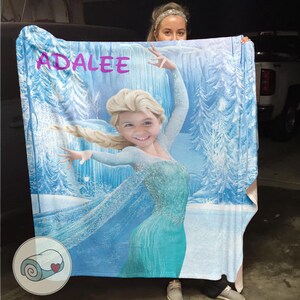 May include: A light blue blanket featuring a cartoon image of a girl dressed as a princess, with the name "ADALEE" in pink letters. The background depicts a snowy forest scene. The blanket is made of soft material.