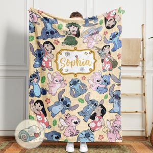 Pu&ograve; includere: Una coperta beige con un motivo ripetuto di personaggi dei cartoni animati in blu e rosa. La coperta ha un disegno centrale con il nome "Sophia" in oro all'interno di una cornice decorativa. La coperta &egrave; realizzata in un materiale morbido.