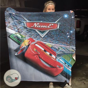 May include: A plush blanket featuring Lightning McQueen from the animated movie Cars. The blanket has a vibrant design with a race track background and the word "Name" in a stylized font. The blanket is primarily red, blue, and green.