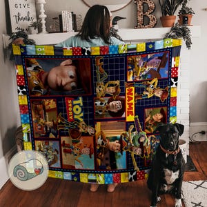 May include: A colorful Toy Story-themed blanket featuring images of Woody and other characters. The blanket has a patchwork design with a blue background and a border of various patterns and colors. The blanket is being held up by a person.