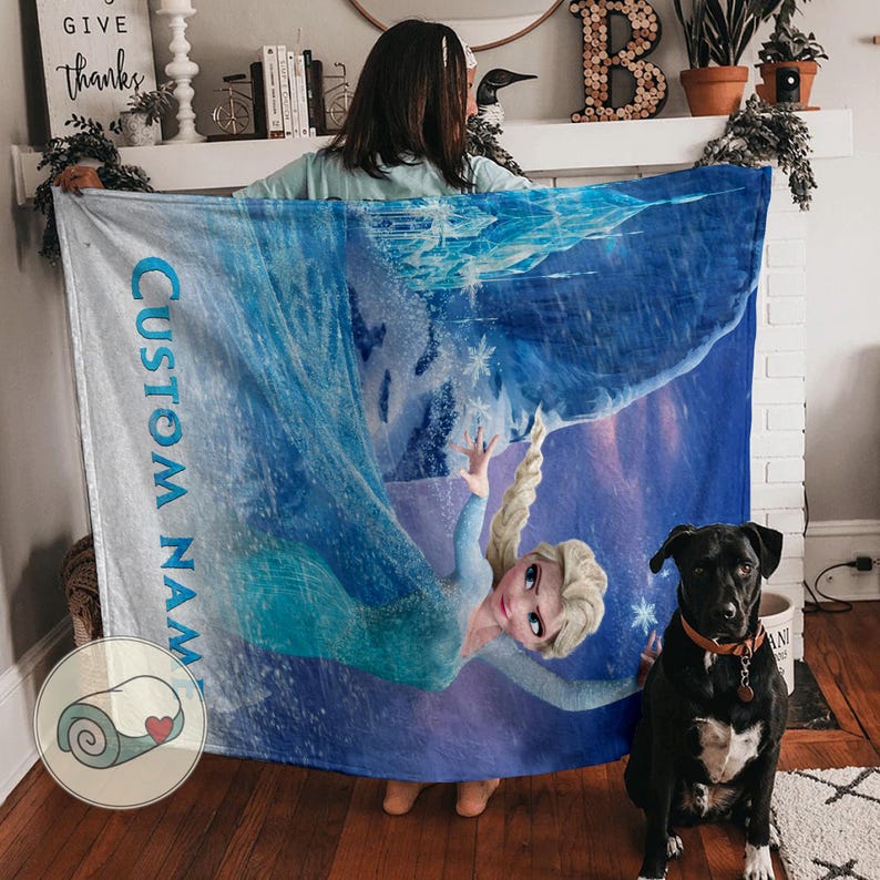 Puede incluir: Una manta azul y blanca con un personaje de dibujos animados. La manta tiene las palabras "CUSTOM NAME" impresas verticalmente en azul. Un perro negro con un collar marr&oacute;n est&aacute; en la imagen.