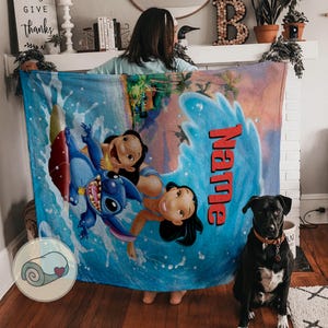 May include: A blue blanket featuring a cartoon illustration of Lilo and Stitch in a wave. The word "Name" is printed in red. A black dog with a brown collar sits in front of the blanket.