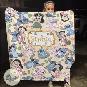 Pu&ograve; includere: Una coperta beige con un motivo ripetuto di personaggi dei cartoni animati, tra cui un alieno blu e una ragazzina. La coperta ha un elemento decorativo centrale con il nome "Sophia" in lettere dorate.