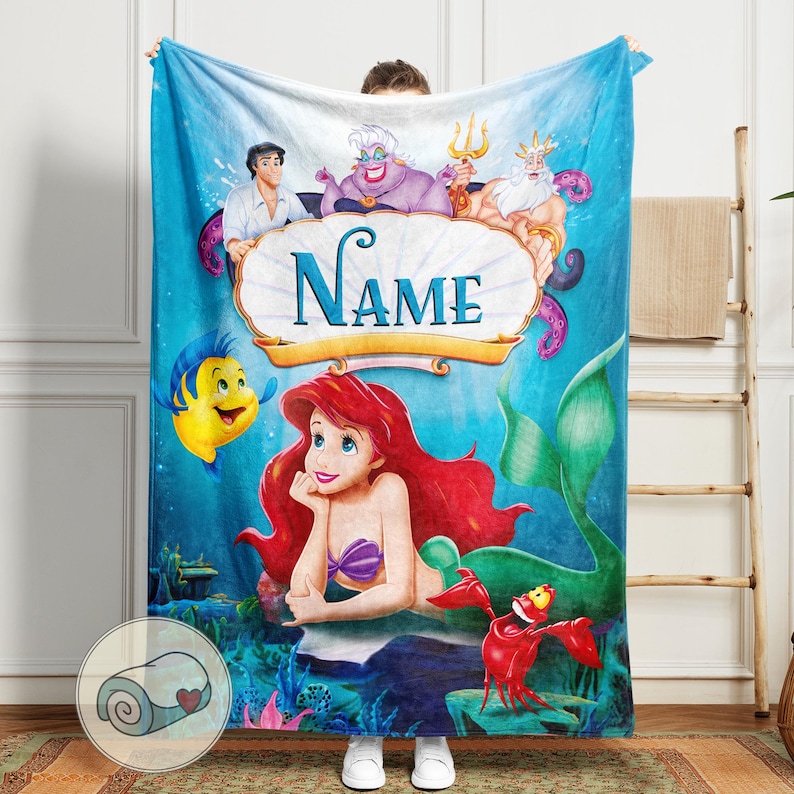 May include: A blue blanket featuring Ariel from The Little Mermaid, with Flounder, Sebastian, and other characters. The blanket has the word "Name" in a decorative font. The background is an underwater scene.