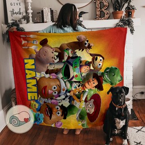 May include: A plush, rectangular blanket featuring characters from the animated film "Toy Story." The blanket has a red border and a gradient background of yellow and orange. The word "NAME" is printed vertically in yellow and blue.