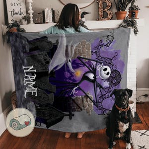 May include: A gray blanket featuring a design with Jack Skellington, a ghost, and a graveyard scene. The blanket has a purple and black color scheme with the word "NAME" printed vertically on the left side.