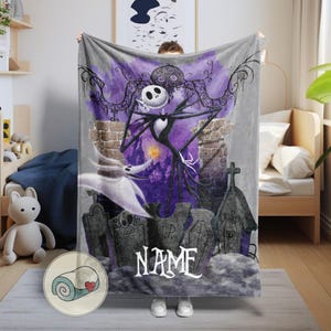 May include: A gray blanket featuring a spooky illustration of Jack Skellington from The Nightmare Before Christmas. The design includes a purple background, tombstones, and the word "NAME" in white. The blanket is held up by a person.