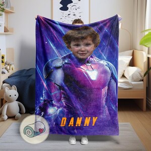 Op de afbeelding: Een gepersonaliseerde pluche deken met het gezicht van een kind bovenop een Iron Man-pakontwerp. De deken is overwegend paars met blauwe en witte accenten, en de naam "DANNY" is in oranje gedrukt.