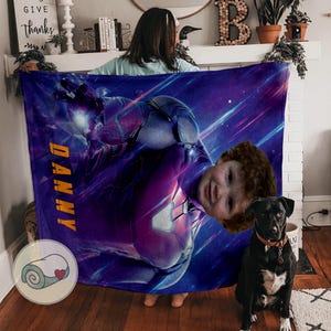 Op de afbeelding: Een gepersonaliseerde deken met een superheldenontwerp en de naam "DANNY" in het geel. De deken heeft een paarse en blauwe ruimte-thema achtergrond. Het gezicht van een kind is op het lichaam van de superheld geplaatst.