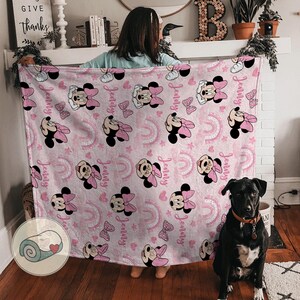 May include: A pink blanket featuring a pattern of Minnie Mouse, bows, rainbows, and the name "Jenny" in a repeating design. The blanket is held up by a person, with a black dog sitting in front of it.