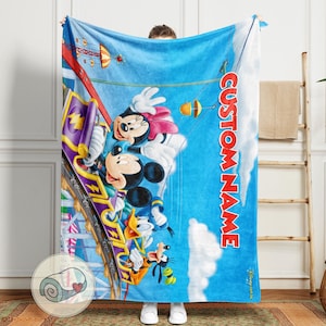 May include: A blue blanket featuring Mickey Mouse, Minnie Mouse, Donald Duck, and Goofy on a roller coaster. The blanket has a blue sky background with white clouds and the text "CUSTOM NAME" in red.