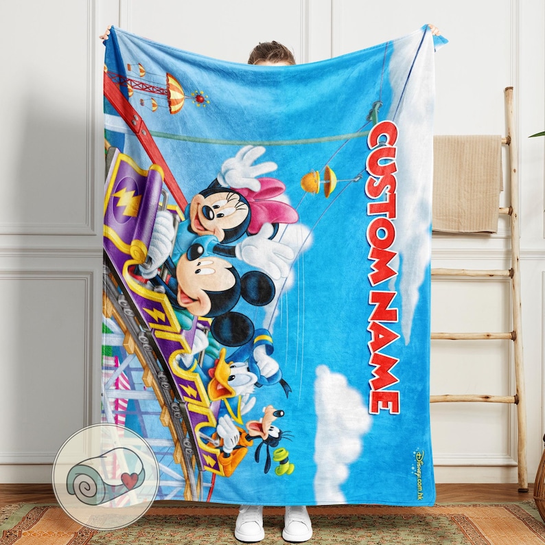 May include: A blue blanket featuring Mickey Mouse, Minnie Mouse, Donald Duck, and Goofy on a roller coaster. The blanket has a blue sky background with white clouds and the text "CUSTOM NAME" in red.