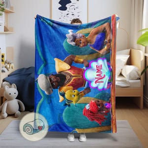 May include: A blue blanket with cartoon characters. The blanket has a repeating teal pattern. Characters include a man in red and gold, a woman in purple, and a mermaid with red hair. A seashell shape has the word "Name".