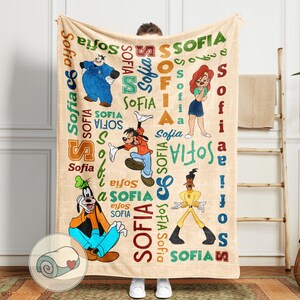 May include: A beige blanket featuring cartoon characters and the name "Sofia" in various colors and fonts. The blanket includes images of Goofy, Pete, and other characters. The word "Sofia" is repeated throughout the design.