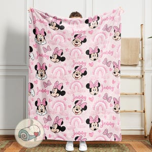 May include: A pink blanket featuring a repeating pattern of Minnie Mouse, bows, rainbows, hearts, and stars. The name "Jenny" is written throughout the design. The blanket is held up by a person.