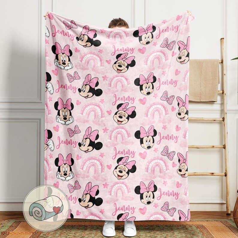 May include: A pink blanket featuring a repeating pattern of Minnie Mouse, bows, rainbows, hearts, and stars. The name "Jenny" is written throughout the design. The blanket is held up by a person.