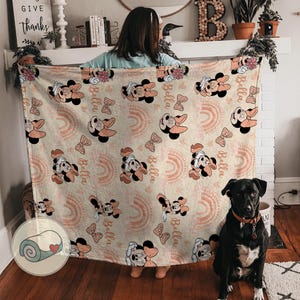 May include: A beige blanket featuring a repeating pattern of Minnie Mouse, bows, and rainbows in shades of peach and pink. The name "Bella" is also printed on the blanket. The blanket is displayed indoors.