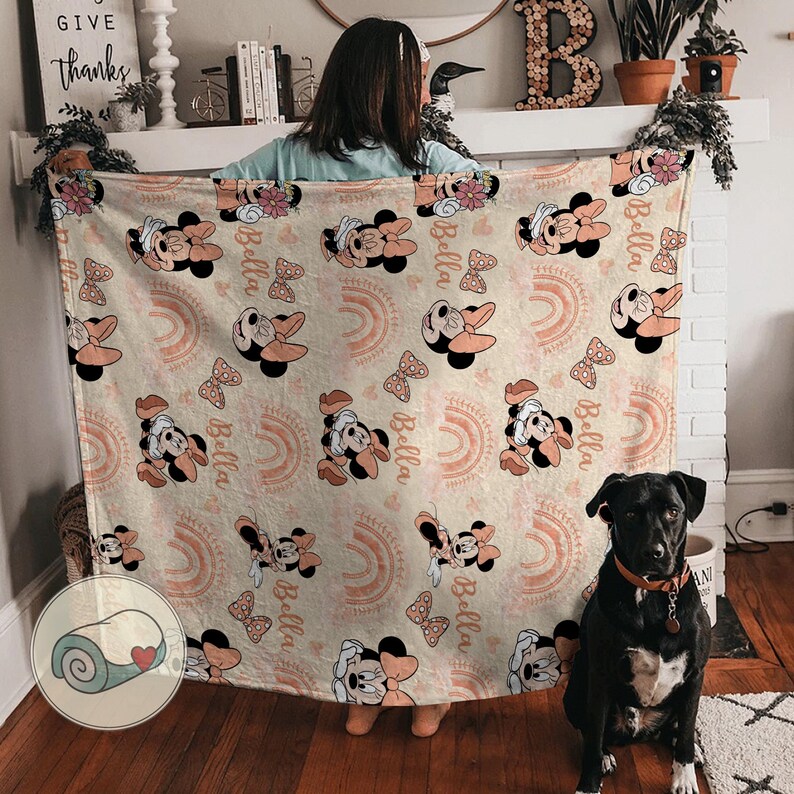 May include: A beige blanket featuring a repeating pattern of Minnie Mouse, bows, and rainbows in shades of peach and pink. The name "Bella" is also printed on the blanket. The blanket is displayed indoors.