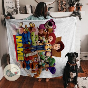 May include: A white blanket with a vibrant collage of Toy Story characters. The blanket has a red and yellow vertical stripe with the word "NAME". A black dog with a brown collar sits on a wooden floor.