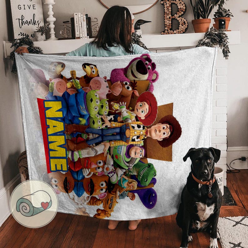 May include: A white blanket with a vibrant collage of Toy Story characters. The blanket has a red and yellow vertical stripe with the word "NAME". A black dog with a brown collar sits on a wooden floor.