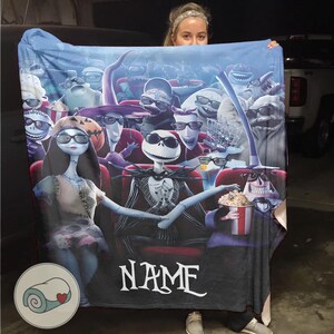 Puede incluir: Una manta azul con una escena de pel&iacute;cula con personajes de The Nightmare Before Christmas. La manta muestra a Jack Skellington y Sally en un cine, rodeados de otros personajes. La palabra "NAME" est&aacute; impresa en la manta.