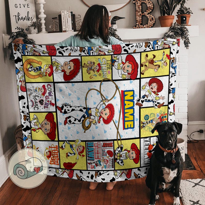 Op de afbeelding: Een wit en gele deken met een cartoon cowgirl ontwerp en het woord "NAME" in blauw en geel. De deken heeft een koeienprint rand en diverse cartoon afbeeldingen. Een zwarte hond zit voor de deken.