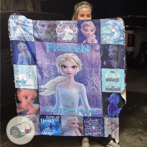 May include: A custom-name Frozen-themed blanket featuring Elsa. The blanket has a purple and blue color scheme with images of Elsa and quotes like "Fear is What" and "The Cold Never Bothered Me Anyway".