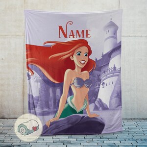 May include: A lavender blanket featuring a cartoon illustration of a mermaid with red hair, a silver seashell bra, and a green tail. The word "Name" is printed in red script above the mermaid. A castle is in the background.