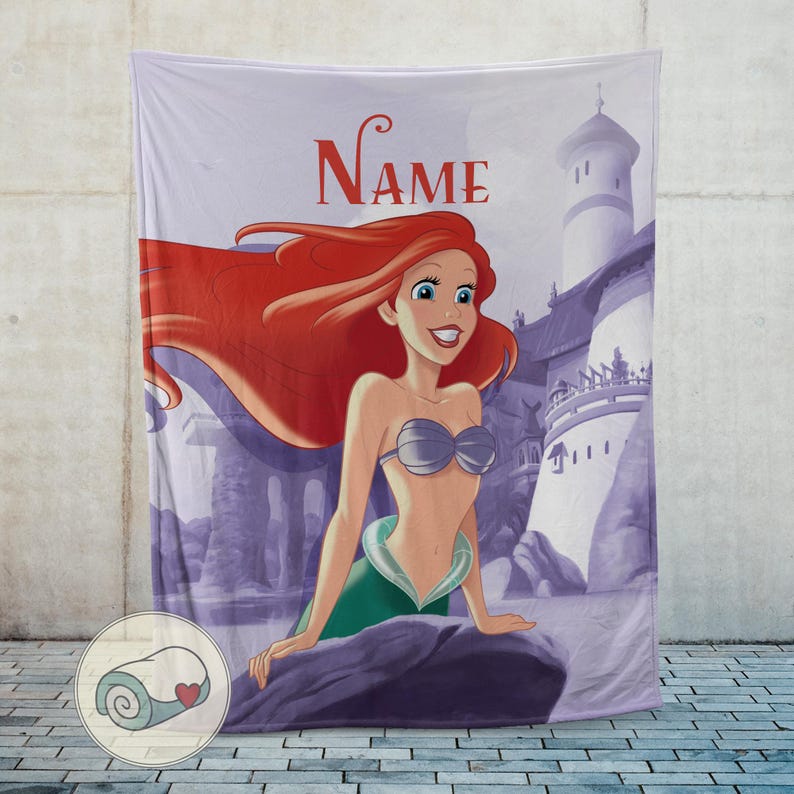 May include: A lavender blanket featuring a cartoon illustration of a mermaid with red hair, a silver seashell bra, and a green tail. The word "Name" is printed in red script above the mermaid. A castle is in the background.