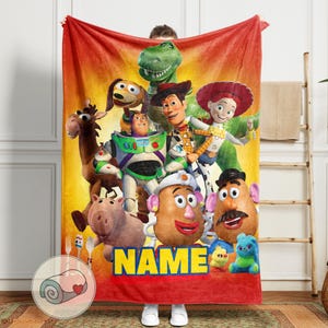 May include: A red and yellow blanket featuring a group of animated characters. The blanket has a vibrant design with characters like Woody, Buzz Lightyear, and Mr. Potato Head. The word "NAME" is displayed in yellow block letters.
