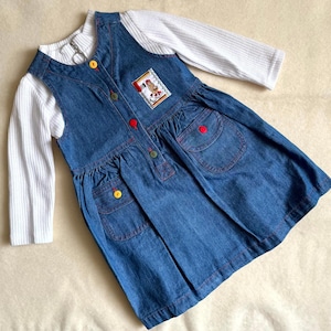 May include: A blue denim pinafore dress layered over a white long-sleeve ribbed top. The dress features a front button fastening with multicoloured buttons, two front pockets with button flaps, and a small rectangular patch with a graphic design.