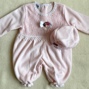 May include: A light pink baby romper and matching hat set. The romper features a lace bodice, white lace trim, and a bow with floral accents. The hat has a matching bow and lace trim. The set is made of a soft, plush material.