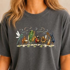May include: A charcoal grey short-sleeve t-shirt featuring a graphic illustration of six insects playing musical instruments, including a dragonfly, a beetle on drums, a cicada on saxophone, a ladybug on guitar, a bee, and a beetle on a harp.