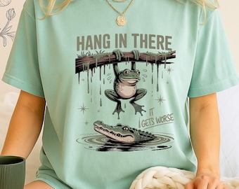 May include: A pale green t-shirt featuring a cartoon frog hanging from a branch, with an alligator below. The text "HANG IN THERE" and "IT GETS WORSE" are printed on the shirt. The shirt is a "Chalky Mint" colour.