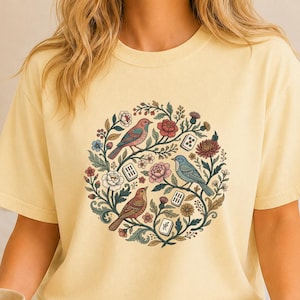 May include: A pale yellow short-sleeve t-shirt featuring a circular graphic print of three birds, various flowers, and small rectangular tiles. The design includes red, blue, and green botanical elements arranged in a wreath-like pattern.