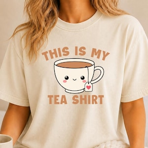 May include: An ivory short-sleeve t-shirt featuring a graphic of a smiling tea cup with a tea bag tag showing a small red heart. The text above the cup reads THIS IS MY and the text below the cup reads TEA SHIRT in a brown sans-serif font.