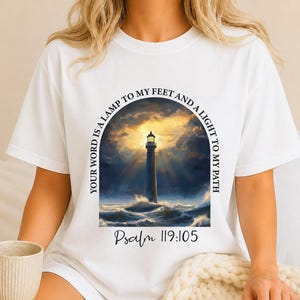 May include: A white short-sleeve t-shirt featuring a graphic of a lighthouse amidst a stormy sea at sunset. The text reads, "YOUR WORD IS A LAMP TO MY FEET AND A LIGHT TO MY PATH" in an arch, with "Psalm 119:105" printed below the image.