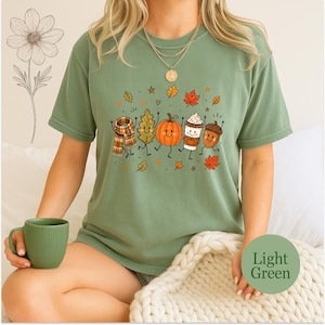May include: A light green short-sleeve t-shirt featuring a graphic print of a scarf, an oak leaf, a pumpkin, a coffee cup, and an acorn, all with smiling faces and thin black limbs, surrounded by small autumn leaves and stars.