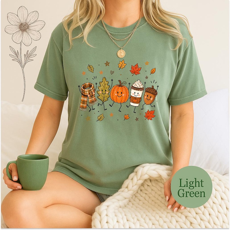 May include: A light green short-sleeve t-shirt featuring a graphic print of a scarf, an oak leaf, a pumpkin, a coffee cup, and an acorn, all with smiling faces and thin black limbs, surrounded by small autumn leaves and stars.