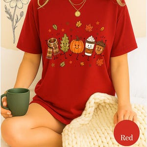 May include: A red short-sleeved t-shirt featuring a graphic print of a scarf, a green leaf, an orange pumpkin, a coffee cup, and an acorn, all with smiling faces and limbs. The design includes small scattered autumn leaves and stars.