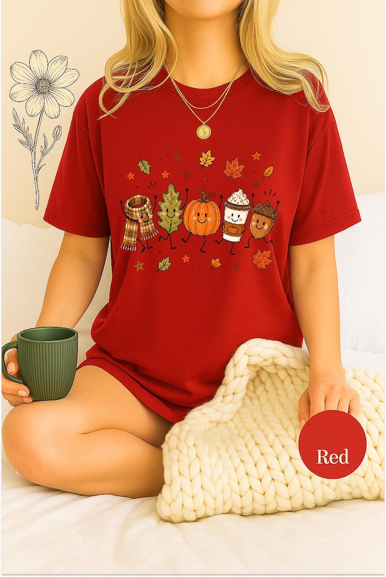 May include: A red short-sleeved t-shirt featuring a graphic print of a scarf, a green leaf, an orange pumpkin, a coffee cup, and an acorn, all with smiling faces and limbs. The design includes small scattered autumn leaves and stars.