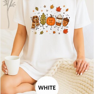 May include: A white short-sleeved t-shirt featuring a colorful graphic print of anthropomorphic autumn elements including a plaid scarf, a green leaf, an orange pumpkin, a coffee cup, and an acorn, all surrounded by small falling leaves and stars.
