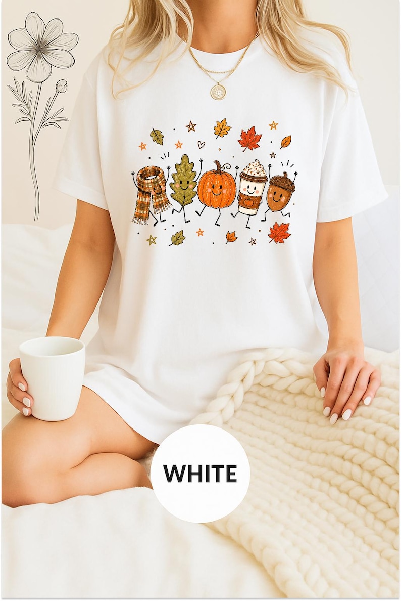 May include: A white short-sleeved t-shirt featuring a colorful graphic print of anthropomorphic autumn elements including a plaid scarf, a green leaf, an orange pumpkin, a coffee cup, and an acorn, all surrounded by small falling leaves and stars.