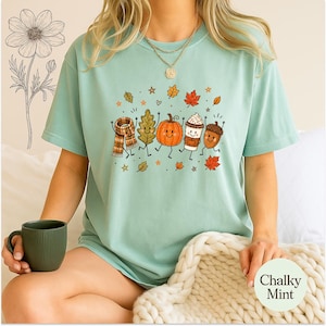 May include: A mint green short-sleeve t-shirt featuring a graphic print of a plaid scarf, a green leaf, an orange pumpkin, a coffee cup, and an acorn, all with smiling faces, surrounded by small autumn leaves and stars on the front.