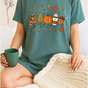 May include: A blue-green short-sleeve t-shirt featuring a graphic print of a smiling scarf, leaf, pumpkin, coffee cup, and acorn dancing together, surrounded by small autumn leaves and stars. The shirt has a crew neckline and a relaxed fit.